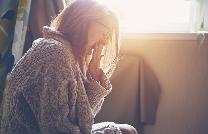 Woman wearing glasses and grey sweater sitting on the floor and yawning; bright stream sunlight coming through the window